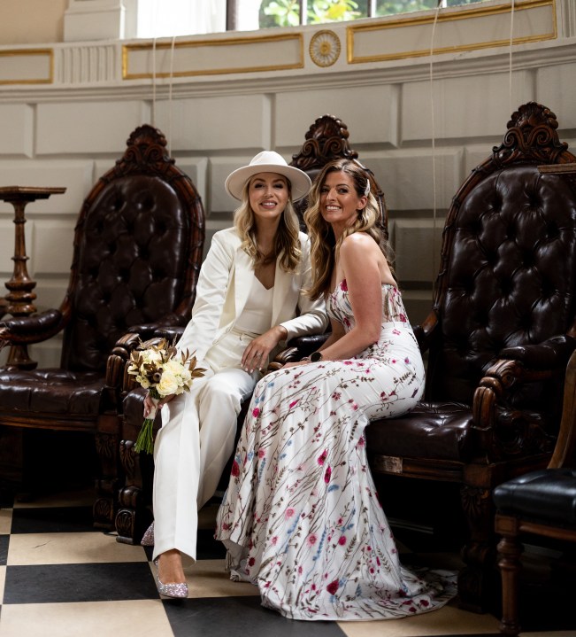Two brides in wedding attire sitting in big black chairs in Trinity's public theatre