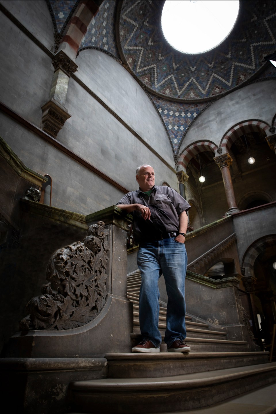 Patrick Wyse Jackson, Professor in Geology standing on the staircase of the Museum Building at Trinity, wearing a grey t shirt and blue jeans