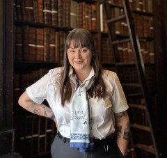 Library Guard Shauna in the Old Library, wearing black trousers, a belt, tie, and white shirt
