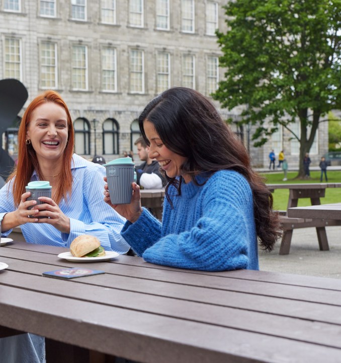Two women at a picnic table on Trinity campus, each holding a coffee cup, sharing a moment together in a peaceful outdoor environment.