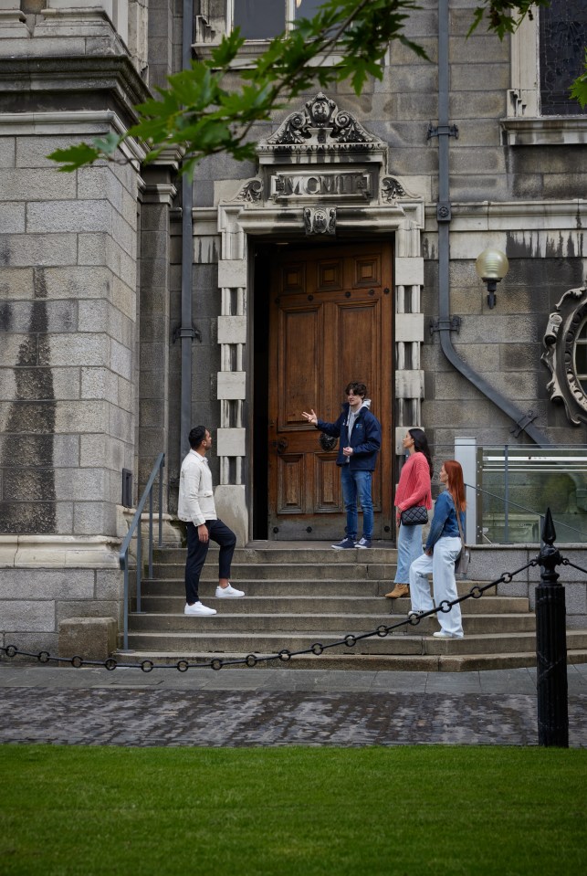 A Trinity tour guide standing on the steps of the Graduate Memorial Building with three tourists during their walking tour of Trinity College in Dublin.