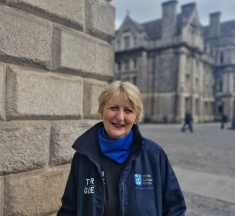 Image of Anne standing outside on Trinity campus, wearing a blue snood and navy Trinity Guide jacket