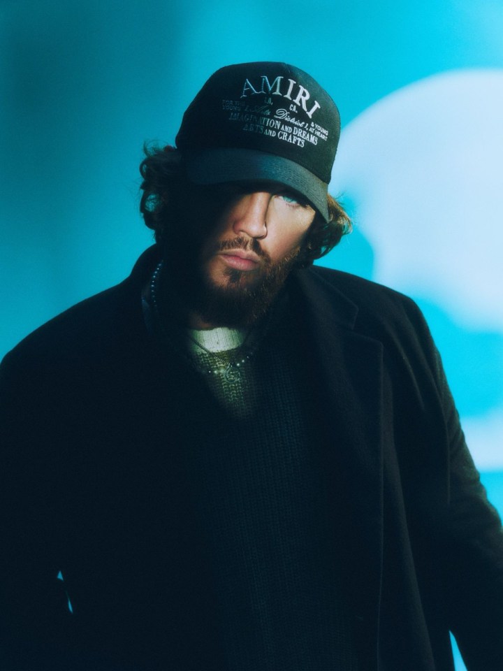 Image of Singer James Arthur in half shadow, half light, wearing dark clothing and a cap that says โAmiriโ.