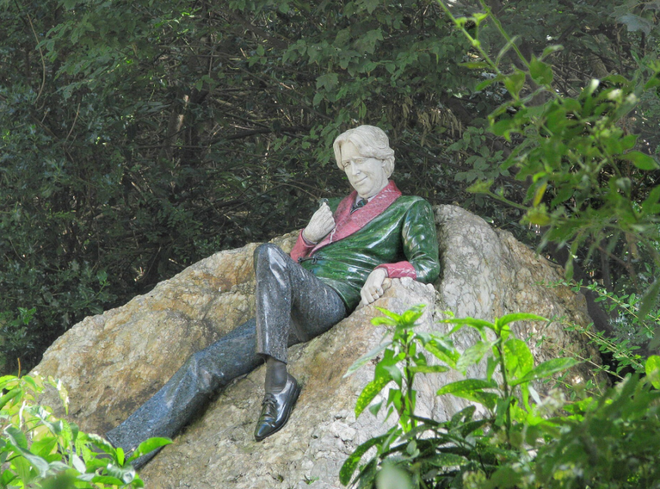 A sculpture of a man wearing a green and pink coat and black trousers, lying on a rock.